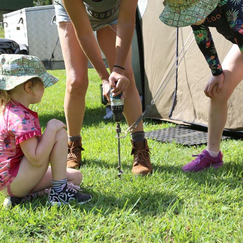 Susu using a power drill to drive a GroundGrabba Pro 300mm screw-in tent peg into the ground while her granddaughters watch, showing easy installation for camping setups.