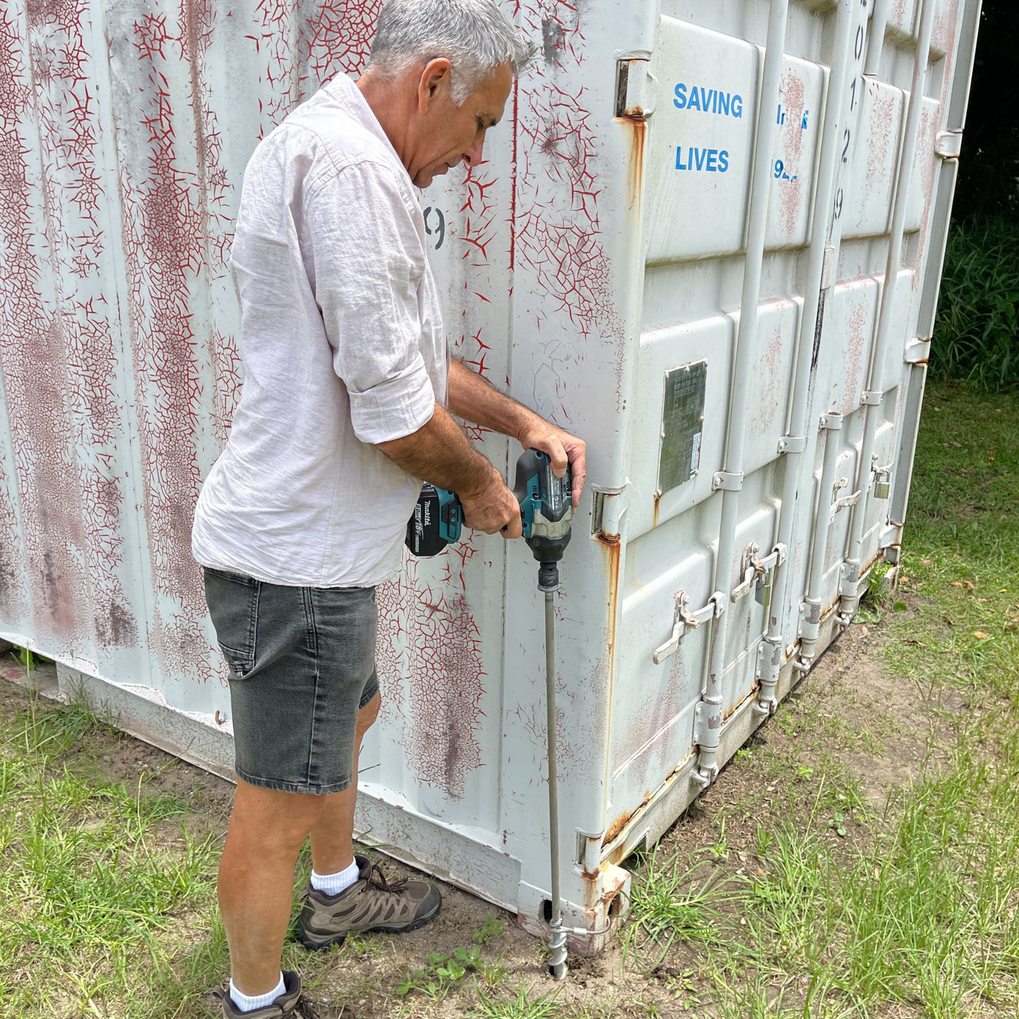 Person drilling steel ground anchor with steel cable looped through shipping container anchor point.