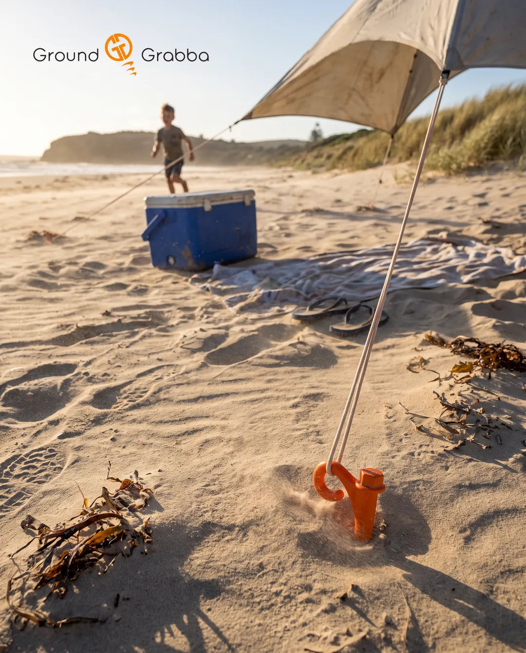 GroundGrabba Lite screw-in sand peg installed flush in beach sand, guy rope through the built-in hook holding a canvas beach shelter in the wind